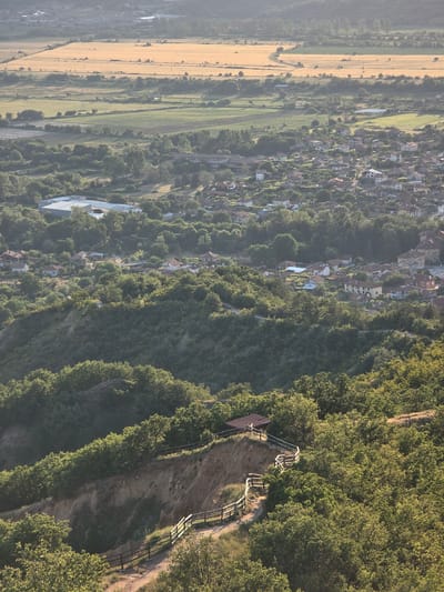 The image displays an aerial perspective of the landscape surrounding Stob, Bulgaria, characterized by natural and developed elements under hazy, diffused sunlight.
In the foreground, a steep, densely forested hillside slopes downwards from the right towards the lower left. The hillside is covered with numerous deciduous trees, primarily light green at their canopy tops where sunlight hits, transitioning to darker green in shaded areas. A dirt path, featuring a rustic wooden fence along its outer edge, curves down the slope. Exposed reddish-brown earth is visible where the path is cut into the terrain, suggesting a steep drop-off. A small, dark-roofed shelter or viewing platform is positioned on a level section of the path near the upper-right foreground, surrounded by trees and overlooking the valley below.
The midground features a rural town or village interspersed with extensive tree cover. Numerous houses with tiled, predominantly reddish-brown roofs are visible, along with some buildings featuring lighter grey or white roofs. Several larger, light-colored industrial or commercial structures with expansive roofs are located on the western outskirts of the town. Roads and smaller pathways wind through the settlement.
In the background, the landscape transitions into large agricultural fields, extending towards a distant, hazy horizon. A prominent golden-yellow field, likely indicating mature crops, occupies a significant portion of the upper background. Adjacent fields appear green. Scattered trees and tree lines define the edges of these fields and follow the contours of the distant terrain. No individuals or distinct human activities are discernible within the frame.