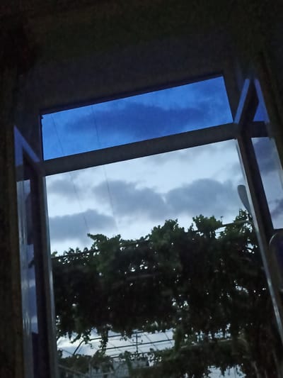 The image is a view through an open window looking out at a cloudy sky and foliage. The upper part of the window frame is dark, suggesting it is indoors. The sky is a mix of dark and light grey clouds, with a strip of brighter blue sky visible at the top. Below the sky, there is a dense mass of dark green foliage, possibly trees or vines, with some lighter sky visible through gaps. The overall impression is of an overcast or twilight scene. The location is identified as Chust, Uzbekistan.