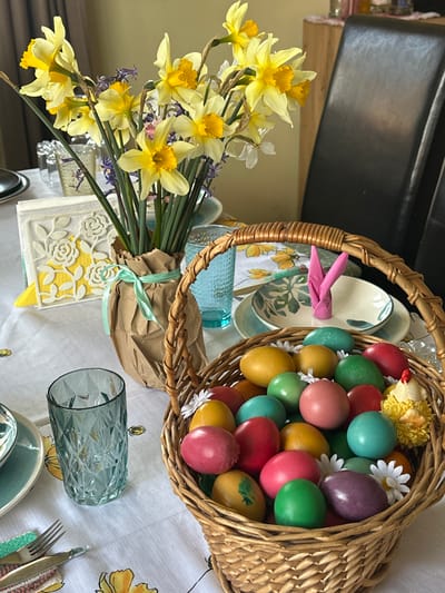 A still life composition on a table covered with a white tablecloth, featuring a basket filled with colorful Easter eggs. The eggs are various shades of red, green, yellow, pink, and turquoise. Small white daisy-like flowers are scattered among the eggs. Next to the basket is a bouquet of yellow daffodils wrapped in brown paper and tied with a light green ribbon. Two blue faceted glasses are also on the table. Plates with floral patterns and cutlery are visible on the edges of the table. A pink bunny-shaped object is placed near the basket. A decorative white card with a floral cutout design and a yellow backing is also visible. The background includes a black countertop. The location is not visible in the image, so it can't be confirmed if it is in the city of Bryastovets, Bulgaria.