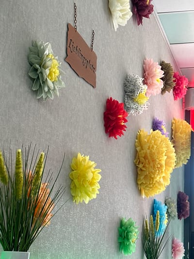 The image depicts a wall decorated with various artificial flowers and a metal sign. The sign, shaped like a stylized plaque, is hung by two chains and features the text "Lieblingsplatz" cut out in a decorative font. The flowers are made of paper or similar material and are arranged in clusters and individually across the wall. Colors include light green, yellow, red, pink, purple, blue, and maroon. In the lower left and lower right of the image, there are arrangements of artificial grass-like plants in white planters, with an orange flower partially visible in the lower left. The wall itself has a textured, light gray surface. The lighting suggests an indoor environment, possibly a public space or a decorated wall in a home or business. Without further context or external information, the specific location in Falkensee, Germany cannot be determined from the image alone.