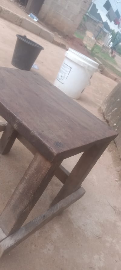 In a daytime outdoor setting in Bende, Nigeria, a handmade, dark wood, four-legged stool sits on a rough, tan concrete surface. Behind the stool are a black plastic bucket, a white plastic bucket, and an unidentified concrete structure. In the far background, a building under construction and clothes hanging on a line are visible. No people are near the stool.