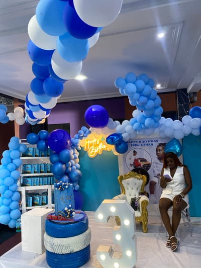 The image shows a birthday party for a child named Joel-Ethan, who is turning three years old. The party is decorated with blue and white balloons, arranged in arches around the room. There is a large, light-up number "3" on a table, and a cake with blue and white frosting. In the background, a wall is decorated with a large, blue and white banner that reads "Happy Birthday Joel-Ethan." A young woman is sitting on a white chair, looking at her phone. She is wearing a white vest and white shorts. There are gift boxes on shelves behind the balloons. The scene is likely at a venue in Bonadikombo, Cameroon.