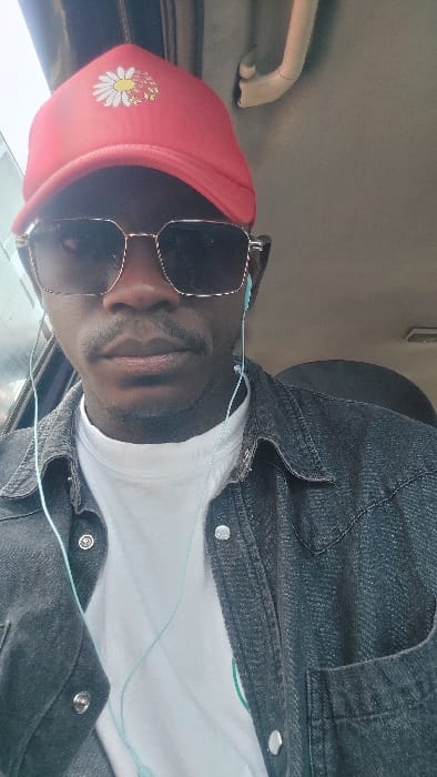 A male individual is depicted in a close-up portrait, seated inside what appears to be a vehicle in the city of Hoss, Nigeria. He wears a red baseball cap adorned with a white daisy design, dark rectangular sunglasses with gold-colored frames, and light blue wired in-ear headphones. His attire consists of a white t-shirt layered under an unbuttoned dark denim-style jacket with silver snap buttons. A mustache is visible above his upper lip. The interior background shows a light-colored headliner, a beige grab handle, and ambient light entering from a window on the left.