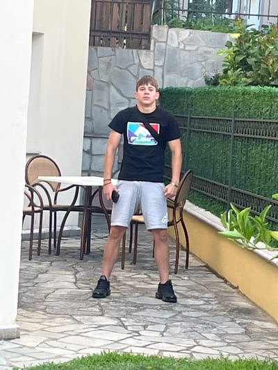 A young male is standing on a stone-tiled patio in front of a building with a green hedge and a metal fence. He is wearing a black t-shirt with a colorful graphic on the front, light gray shorts with an Adidas logo, and black athletic shoes. His left hand is holding a dark-colored smartphone. To his left, there is a round white table with several chairs around it. The background consists of a stone wall and foliage.