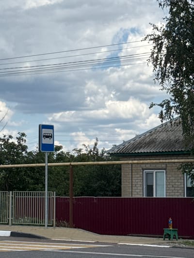 The image shows a street scene in Shulhynka, Ukraine, with a bus stop sign prominently displayed on the left. The sign is blue with a white silhouette of a bus. Behind the sign, there are trees and a brick house with a corrugated roof. A maroon fence runs along the front of the property. In the foreground, a pedestrian crossing is visible on the asphalt road. The sky is cloudy with a mix of grey and white clouds. Power lines are strung across the upper portion of the image. A small green stool with a bottle on it is placed near the fence on the right side of the image. There are no people visible in the scene.