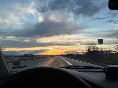 The image shows a view from inside a vehicle driving on a highway, likely in the vicinity of Georgi-Dobrevo, Bulgaria, given the presence of a Shell gas station sign. The road stretches towards the horizon where the sun is setting, creating a bright glare. The sky is filled with clouds. The interior of the vehicle is visible at the top of the frame.