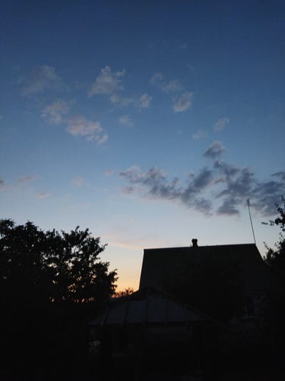 The image depicts a silhouetted view of a rural landscape at dusk, likely in the Кіраўскі сельскі Савет, Belarus. The sky transitions from a deep blue at the top to lighter shades of blue and a pale orange glow near the horizon, indicating the setting sun. Scattered clouds are visible in the sky, some appearing wispy and others more defined. In the foreground, the dark silhouette of trees frames the left side of the image. A house with a visible chimney is silhouetted in the mid-ground, with a darker structure, possibly a greenhouse or shed, in front of it. A thin, tall antenna stands to the right of the house. The overall scene is dark, with the details of the structures and foliage obscured by shadow.