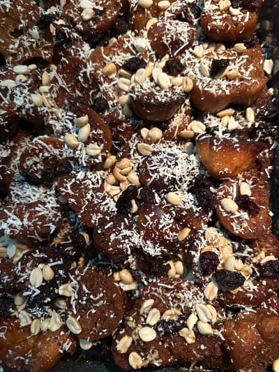 This image depicts a close-up, overhead view of numerous irregularly shaped, deep-fried or baked dough pieces, ranging from golden-brown to dark brown, arranged in a dark-colored tray or pan. Each piece, approximately 2 to 5 cm in diameter, exhibits a glossy, syrupy coating. The items are liberally and unevenly topped with shredded white coconut flakes, light-brown split and whole peanuts, and dark-brown shriveled raisins. The container is filled to capacity with the food. The scene is located in the city of Tres Valles, Mexico. No individuals or specific actions are observed.