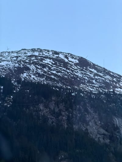 The image displays a majestic mountain face, steeply rising and partially covered in snow against a clear, light blue sky. The mountain dominates the right side of the frame, revealing rugged rock formations interspersed with white patches of snow, suggesting cold weather conditions.

Near the mountain's summit, two slender, mast-like structures, likely communication antennas or towers, are visible, extending into the sky. Further down the mountain face, a series of thin lines or cables, possibly power lines or part of a cable car system, crisscross the rocky terrain. In the bottom left corner, a portion of a large, blade-like structure, potentially from a wind turbine or similar infrastructure, can be faintly seen.

The sky is bright and uniformly light blue, indicating it is daytime. There are no people or discernible animal activities. The scene captures a grand natural landscape featuring prominent human-made installations, characteristic of a mountainous region such as Göschenen, Switzerland.