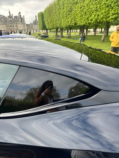 The image shows the rear side of a black car parked in front of the Chateau de Chambord, a large, historic castle in Chambord, France. The car is facing the castle, which is visible in the background. The car's windows are tinted, and the reflection of a woman taking a selfie with a phone is visible in the rear window. The woman is wearing a white shirt and a brown jacket. The car is parked on a grassy field, with a line of trees behind the car. The trees are all green and leafy, and the sky is cloudy. There are a few people in the background, including one man in a yellow jacket. The coordinates of the Chateau de Chambord are approximately 47.616°N, 1.519°E. The image is shot at a slight upward angle. The car is in focus, with a soft focus on the background. The image is composed in a way that emphasizes the car and its reflection. The image is shot in natural light.