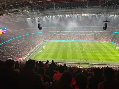 The image shows a view from the stands of Wembley Stadium in London, England, where a football match is taking place. The field is green and surrounded by white lines, with a scoreboard at one end. There are about 20 players on the field, wearing white and dark uniforms. The stands are packed with spectators. The stadium is illuminated by spotlights, and the sky is dark, suggesting it is nighttime. The image is taken from a high vantage point, providing a panoramic view of the stadium and the surrounding area. The coordinates provided place the stadium in Wembley Park, London, England.