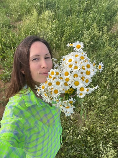 A woman with long, dark brown hair and fair skin is holding a large bouquet of white daisies with yellow centers. She is wearing a green and blue plaid shirt. She is looking at the camera with a slight smile. The background is a field of green grass and wildflowers. The scene is outdoors. The location is described as Пигозино, Russia.