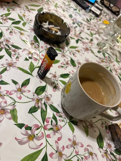 A white mug filled with coffee sits on a table with a floral tablecloth. An ashtray filled with cigarette butts and a black and orange "Turbo" lighter are nearby. A box of what appears to be Chesterfield cigarettes is partially visible in the upper right corner. Glasses and other objects are out of focus in the upper left corner, completing the scene of a casual, lived-in space.