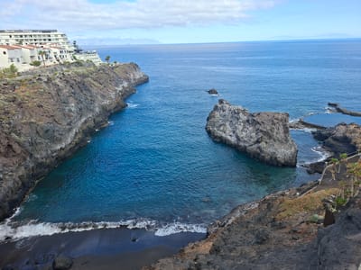 The image depicts a coastal view in Santiago del Teide, Spain, featuring a bay with dark volcanic sand and clear blue water. On the left, a rocky cliff rises, topped by a large white hotel building with the "Barceló Santiago" sign visible. The hotel has multiple stories and balconies, with some greenery around it. To the right of the bay, a natural rock formation juts out into the sea. Further to the right, a man-made saltwater swimming pool with a curved edge is visible, built into the rocky coastline. The sea stretches to the horizon, with faint mountain ranges visible in the distance. The sky is partly cloudy with white clouds against a blue background. The overall scene is one of a coastal resort area with a mix of natural rock formations and developed infrastructure.