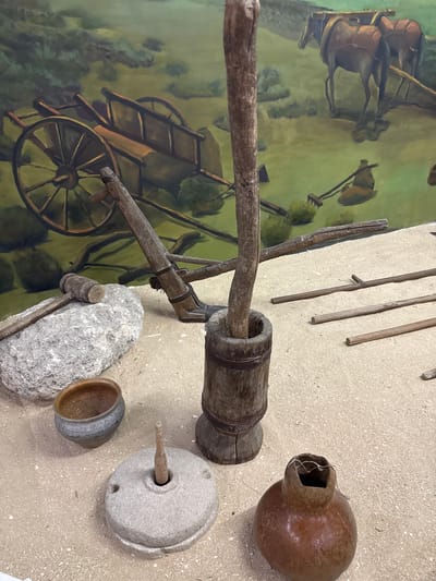 The image displays a diorama of historical agricultural tools and artifacts set against a painted mural depicting a pastoral scene. In the foreground, on a sandy surface, rest several objects: a large, grey, circular grinding stone with a wooden pestle inserted in its center; a small, dark, rounded pot; a wooden mallet resting on a large, rough stone; a wooden mortar with a wooden pole inserted into it; and a brown, gourd-like vessel with a narrow opening. Several long, wooden poles are scattered on the sand behind these items. The background mural shows two horses harnessed to a wooden plow, and a wooden cart with large spoked wheels, all set in a green, hilly landscape. The overall impression is that of an exhibit intended to showcase traditional farming methods. The location is identified as Fort-Şevtşenko, Kazakhstan.