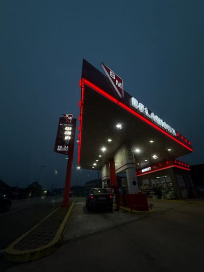 The image captures a gas station at night or late dusk, illuminated by artificial lights against a dark blue sky. The scene is set in Brčko, Bosnia and Herzegovina, specifically at a "BELAMIONIX" station, as indicated by prominent signage.
The main subject is the gas station canopy, which features a distinctive triangular shape and is outlined with bright red LED strip lighting. The "BELAMIONIX" name is displayed in large, white, illuminated letters along the edge of the canopy, accompanied by a smaller "BM" logo. Beneath the canopy, multiple bright lights illuminate the area around the pumps.
To the left of the main structure, a tall, red pole supports an electronic price display board. This board also features the "BM" logo and "BELAMIONIX" text at the top. Below this, three prices are clearly visible: "2.33", "2.33", and "1.23". At the bottom right of the display, "00-24" indicates 24-hour operation.
Under the canopy, several gas pumps are visible, with one dark-colored car parked beside a pump, suggesting it is either fueling or waiting to fuel. To the right of the pumps, an attached convenience store or "BM MARKET" is brightly lit, with its interior visible through large windows, showcasing various products. The "BM MARKET" text is also visible above the store entrance.
The ground consists of a dark asphalt road surface and a patterned brick pathway with a yellow curb, leading towards the foreground. The overall impression is that of a functional, well-lit commercial establishment operating during nighttime hours.