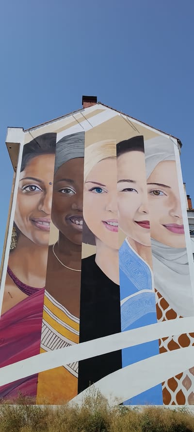 A mural of five women's faces is painted on the side of a building in Siero, Spain. The women are diverse in ethnicity and appearance. The mural is on the facade of a building with a tiled roof and a chimney. The sky is clear and blue. The building is surrounded by dry grass and vegetation at the bottom of the image. The mural is divided into vertical sections, each depicting a portion of a woman's face. From left to right, the first woman has dark hair and is wearing a dark red garment and large gold earrings. The second woman has dark skin and is wearing a grey headscarf and a necklace. The third woman has blonde hair and blue eyes. The fourth woman has dark hair and is wearing a blue garment with a geometric pattern. The fifth woman is wearing a light-colored headscarf and has brown eyes. The mural extends to the bottom of the building, where there is a pattern of brown giraffe-like spots on a light background. White diagonal stripes are painted across the mural.