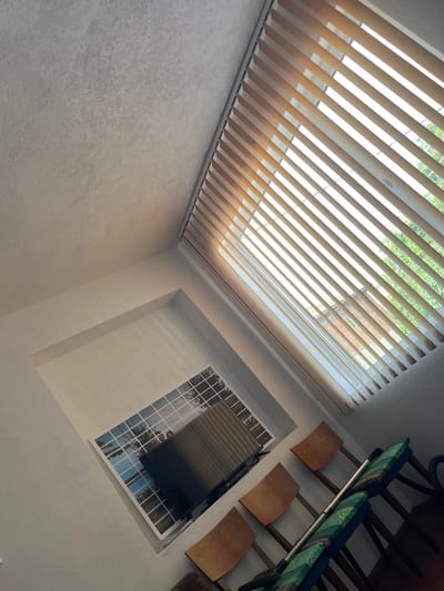 A low-angle interior shot shows a white-walled room with a large window covered by closed vertical blinds through which light streams. A smaller window reflecting a landscape scene is mounted on the adjacent wall. Wooden chairs line the top right of the frame. The location is not discernible within the city of Dalgopol, Bulgaria. There are no people visible.