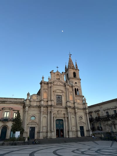 The image captures the Basilica di San Sebastiano in Acireale, Italy, situated against a clear blue sky with a crescent moon visible. The basilica's facade is intricately detailed with baroque architectural elements, including columns, statues, and ornate window surrounds. A portion of a neighboring building is visible on the right side of the frame, as is a patterned stone pavement. A bicycle is parked near the building at street level. The perspective is from below, emphasizing the height and grandeur of the basilica.