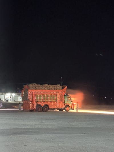 An ornate, brightly painted truck, likely a Pakistani truck, is parked at night on a dirt or gravel lot. The truck is laden with numerous sacks, possibly containing grain or other goods, piled high in its bed. The truck is red and green, with intricate, colorful decorations along its sides. A single headlight on the truck illuminates the immediate area, casting a warm glow and creating a cloud of dust or exhaust behind it.
In the background, to the left of the truck, there appears to be a small building or structure with lights on, suggesting a roadside establishment or dwelling. A few figures are visible near this structure, one of whom is walking away from the truck. To the right of the truck, a man in a light-colored outfit stands facing the truck, illuminated by the truck's light. The environment is dark, indicating it is nighttime. The ground is a light-colored, coarse material. The sky is dark with some faint, distant lights visible.