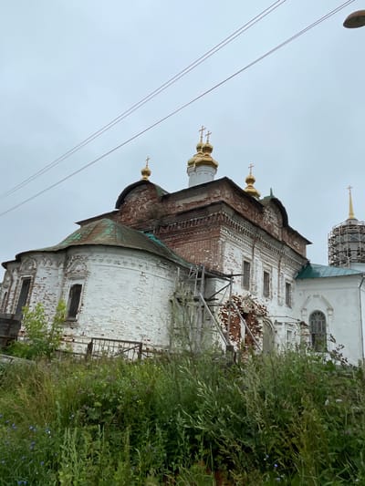 An outdoor, low-angle, medium shot shows an old, white brick church with a red brick upper section, possibly under construction or renovation, in the city of Kstynino, Russia. The church has several golden domes topped with crosses. Scaffolding is visible on the side of the building. The church is surrounded by overgrown green grass and foliage, with a grey, overcast sky above. Power lines are visible in the upper portion of the image.