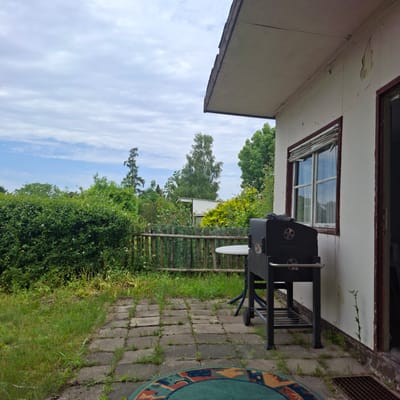 The image depicts a slightly overcast outdoor scene, likely a backyard or patio area, with a white-sided building on the right. In the foreground, a black charcoal grill is positioned on a paved stone patio. To the left of the grill, a small, round white table with blue legs is visible. The patio is bordered by overgrown grass and a wooden fence. Beyond the fence, a lush green hedge runs across the middle ground, with various trees and foliage behind it. In the background, several trees are visible, including a tall, slender tree on the left and a larger, more rounded tree towards the center, with a white, shed-like structure visible behind the fence and hedge. The sky is a pale blue with scattered white clouds. The building on the right shows signs of wear, with peeling paint and a window with blinds. There are no people or animals present in the image. The scene suggests a residential outdoor space in Hermsdorf, Germany, potentially in a garden or yard.