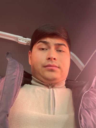The image shows a close-up, chest-up portrait of a young person, appearing male, with dark hair and dark eyes, looking directly at the camera. The image is oriented with the person's head tilted to the right, suggesting it was taken with the camera rotated 90 degrees counter-clockwise.
The person has short, dark hair, neatly styled. Their skin tone is light to medium, and a warm, reddish light source illuminates their face and parts of their clothing, casting a subtle glow. Their expression is neutral, with lips relaxed. They have distinct eyebrows and a faint hint of stubble on their chin.
They are wearing a light-colored, possibly beige or cream, ribbed turtleneck or quarter-zip pullover sweater. A silver zipper is visible down the front of the sweater. Over this, they are wearing a dark-colored (likely black or dark grey/blue) puffer jacket. A faint, lighter-colored circular or crescent-shaped emblem/logo is visible on the upper left arm/shoulder area of the jacket.
The background is mostly dark and out of focus, suggesting an indoor or dimly lit setting. A light-colored, somewhat angular structure, possibly part of a chair, headrest, or vehicle interior, is visible behind the person's head and to their left (when oriented correctly). The lighting gives the scene a warm, slightly intimate ambiance. No discernible text is visible in the image.