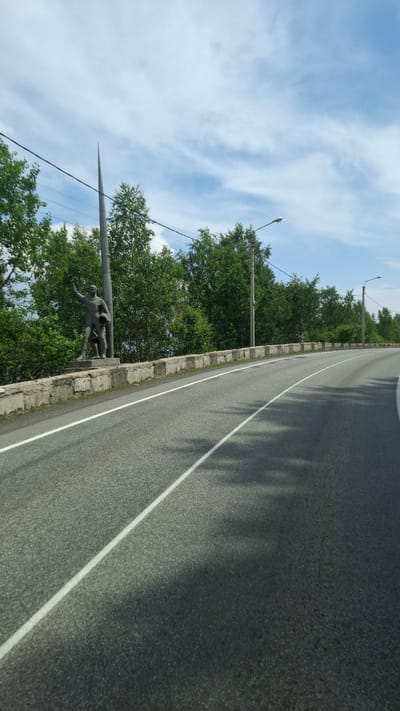 A scenic view of a winding asphalt road in Sukhoy Ruchey, Russia, on a bright, cloudy day. The road is lined with a low concrete barrier, behind which stands a concrete statue of a man holding a helmet, with a smaller figure at his feet. Lush green trees form a dense backdrop along the roadside, with several lampposts visible amidst the foliage. The sky is a mix of blue and white clouds. The image appears to be taken from a moving vehicle, given the perspective and the shadows cast on the road.