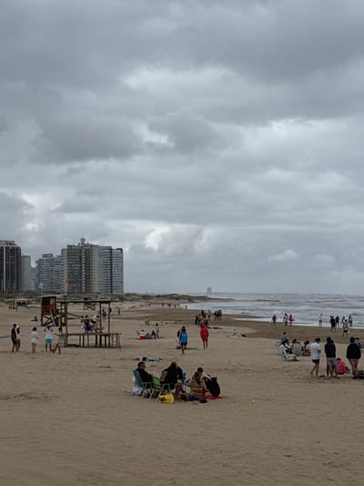This media file depicts a bustling beach scene under an overcast sky, likely in Punta del Este, Uruguay, as indicated by the context. The image is oriented vertically but shows a horizontal expanse of a sandy beach bordered by the ocean on one side and a line of high-rise buildings on the other.
Numerous people are visible across the beach. Many are gathered closer to the water's edge, some standing and observing the waves, which appear moderately rough with whitecaps. Others are scattered further back on the sand, relaxing in beach chairs, under umbrellas (though few are fully open), or simply sitting on towels. Groups of individuals are seen walking or standing, engaging in leisure activities typical of a beach setting. A bright yellow ball is visible on the sand near some seated individuals, hinting at play.
In the midground, a prominent wooden lifeguard stand is positioned on the sand, indicating a supervised beach area. Behind the beach and further in the distance, a series of modern, multi-story buildings line the coast, suggesting an urban or resort environment. The architecture consists of tall, rectangular structures.
The sky is largely covered by thick, grey clouds, indicating an overcast day. This weather condition gives the scene a diffused, soft light, rather than direct sunlight. The overall mood appears relaxed and leisurely, despite the lack of sunshine. No discernible text is visible on signs, clothing, or objects within the image.