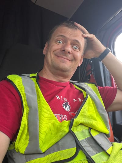 A man with a surprised expression and his hand on his head is captured in a selfie taken from inside a vehicle. He is wearing a red t-shirt with a graphic and text that reads "BIG AP..." and a bright yellow, reflective safety vest over it. His eyes are wide open, and his mouth is slightly open in a smile. The background shows the interior of a vehicle, with dark upholstery and a glimpse of a red seatbelt. The lighting suggests it is daytime. The image does not provide any specific visual cues to determine its location within the city of Chattanooga, United States of America.