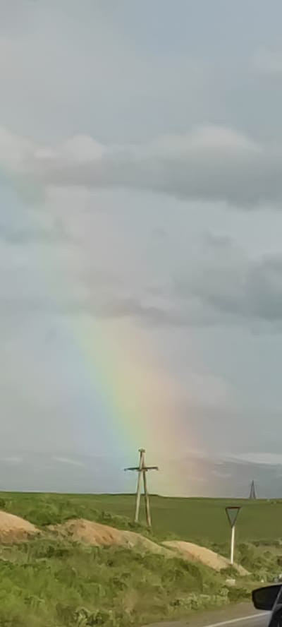 The image shows a landscape near Azatan, Armenia, featuring a partial rainbow in the sky. The landscape includes green fields, small embankments of dirt, a yield sign, and a wooden utility pole in the middle ground. A distant, tall structure is visible on the horizon. Part of a vehicle's interior (specifically, a side mirror) is visible in the lower right corner of the image. The sky is overcast with gray and white clouds.
