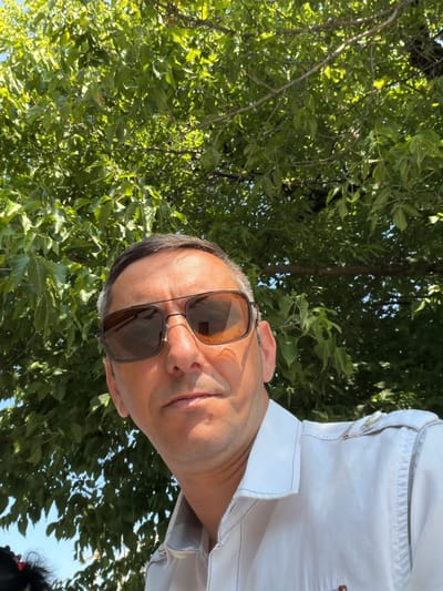 The image is a selfie taken from a low angle outdoors, looking up at a man wearing sunglasses and a light-colored collared shirt. He is positioned in the foreground, with a canopy of green leaves and branches from a tree filling the background. Sunlight illuminates the man's face, casting shadows and highlighting his features. The sky is visible in patches through the foliage. There is no visible indication of the city of Parma, Italy.