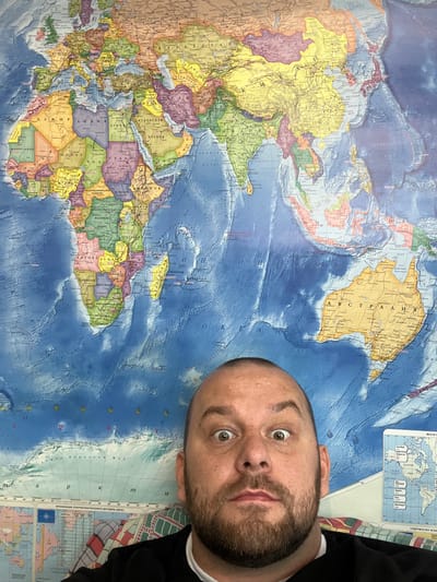 An indoor, vertically oriented close-up shot features a man's head and shoulders positioned over a colorful physical world map. The man, with short dark hair and a beard, looks directly into the camera with wide, slightly surprised eyes and slightly parted lips. He is wearing a dark top with a white collar.

The detailed map, which uses Cyrillic script, covers most of the frame. It appears to be a relief map, showing raised topography for landmasses and texture for ocean depths. The map's orientation is rotated, with North America visible in the upper right, Africa and Europe across the middle, and Australia and Antarctica in the lower left.

Visible text on the map includes country names such as "АВСТРАЛИЯ" (Australia), "КАНАДА" (Canada), "СИРИЯ" (Syria), "ЕГИПЕТ" (Egypt), "АЛЖИР" (Algeria), "НИГЕР" (Niger), "СУДАН" (Sudan), and "ЮЖНЫЙ СУДАН" (South Sudan). Oceanic features like "АТЛАНТИЧЕСКИЙ ОКЕАН" (Atlantic Ocean) and "Земля Королевы Мод" (Queen Maud Land) are also labeled. An inset section displays text related to "ОРГАНИЗАЦИЯ СЕВЕРОАТЛАНТИЧЕСКОГО ДОГОВОРА (НАТО)" (North Atlantic Treaty Organization) alongside a NATO flag, and "СОДРУЖЕСТВО НЕЗАВИСИМЫХ ГОСУДАРСТВ" (Commonwealth of Independent States). The man's face partially obscures parts of the map, notably areas like Greenland and northern Europe. The overall lighting is bright and even.