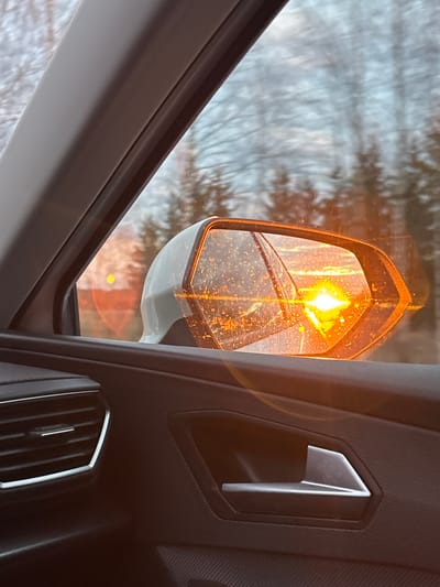 The image presents an interior perspective from a vehicle, with a clear view of the driver's side (or passenger side) external mirror and a portion of the car's dark interior. The central subject is the side mirror, which prominently displays an intensely bright, amber-colored turn signal, actively lit. This light illuminates the surrounding area and reflects within the mirror's glass, creating a radiant glow. The mirror's surface is dotted with numerous small specks, possibly water droplets from rain or melted snow.
Beyond the mirror and through the car window, a blurry outdoor environment is visible. This background consists of indistinct light grey and brown tones, indicative of a natural, possibly wooded setting under overcast skies or during winter, consistent with the location of Печмень, Russia. The car's interior on the right features a dark, textured door panel, a silver-accented door handle, and an air vent with vertical slats. No people or visible text are present. The scene implies the vehicle is either signaling a turn, changing lanes, or is stopped with its hazard lights activated, set against a dim, possibly wintry, outdoor light.