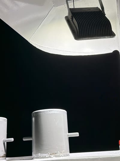 The image shows a close-up view of two distinct objects against a dark background. On the left, a black floodlight with a ribbed surface is mounted on a white, curved structure. The floodlight is angled upwards and illuminated, casting a bright white light onto the surface it's attached to. This white surface appears to be a large, rounded metal component, possibly part of a vessel or structure.

To the right, a cylindrical white object is mounted vertically. It has a metallic sheen and appears to be connected to a larger white structure with visible bolts or fasteners. Two small, cylindrical pegs extend from the top of this white cylinder. This object also shows signs of wear, with some paint chipping.

The scene is set at night, indicated by the dark background and the focused illumination from the floodlight. There are no people visible in the image. The overall impression is one of industrial or structural components, possibly on a ship, bridge, or large outdoor installation. The image has a stark, functional aesthetic, emphasizing the forms and textures of the objects. There is no visible text.