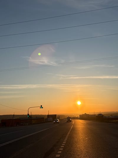 The image depicts a sunrise over a road in Ижевск, Russia. The sky transitions from light blue on the left to orange on the right, where the sun is positioned. Power lines run diagonally across the left side of the frame. A road stretches into the distance, with cars visible. A traffic light with a green light is seen on the side of the road. The road is illuminated by the rising sun, creating a bright reflection. Buildings and trees are visible on the right side of the road.