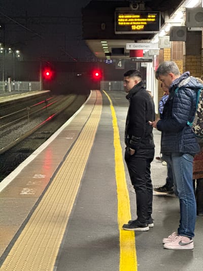The scene depicts a nighttime view of a train station platform at Birmingham International Station, located within the Metropolitan Borough of Solihull, United Kingdom.
On the right side of the image, two male individuals are standing on a paved platform, behind a yellow tactile safety line. The man on the left, of apparent young to middle age, is seen in profile, facing left towards the railway tracks. He has short dark hair with faded sides, is dressed in a black jacket, black trousers, and black sneakers with white soles, with his right hand in his pocket. To his right and slightly behind him stands an older male, wearing a blue quilted jacket, blue jeans, and light pink Nike brand sneakers. He is looking down at a white smartphone held in his hands. A black and white patterned backpack is visible on his back. Partially visible behind these two individuals are the lower legs of at least two other people.
Above the platform, a digital departure board displays "19:40 LONDON EUSTON", with a subsequent line stating "Called at: Arden / Dorridge / Knowle". Below this, it reads "Train is now at Marston Green" and a large number "194307". Beneath the digital board, signs indicate "Gentlemen" and "Waiting room" with directional arrows. The platform itself is constructed of grey asphalt, featuring a continuous yellow tactile safety line. Beyond this, a beige, ridged tactile paving strip runs parallel to the tracks.
To the left, two sets of railway tracks are visible, curving gently into the dark distance. Overhead catenary wires are discernable. Far down the tracks, two prominent red lights, consistent with train signals or tail lights, are visible. The background is dark, illuminated by distant, scattered lights. A small white sign with "2-8" and "4" is visible near the tracks. The station wall on the far right is composed of light-colored bricks, with speakers or announcement devices mounted on it. The overall illumination is provided by artificial overhead lighting on the platform.