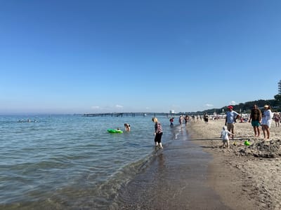 The image depicts a sunny day at the beach in Timmendorfer Strand, Germany. A clear blue sky dominates the upper portion of the frame. In the foreground, the gentle waves of the Baltic Sea lap onto a sandy beach. Several people are visible enjoying the water and the sand. In the sea, some individuals are swimming, while others are wading or playing with a green inflatable ring.
Along the shoreline, a diverse group of people are walking, standing, and playing. A woman in a black and white patterned top and black pants stands in the shallow water, while a woman in a black swimsuit and a child on a green inflatable raft are in the sea nearby. Further down the beach, a family is gathered, with a man in a blue t-shirt and khaki shorts, a man in a green t-shirt and turquoise shorts, and a woman in a white cover-up. A young child in a striped shirt and shorts stands facing away from the camera, seemingly playing with sand toys. In the background, a long pier extends into the sea, with several people walking on it. Trees line the edge of the beach in the distance, and a tall white building is visible to the right of the pier, suggesting a developed coastal resort. The overall scene conveys a lively and pleasant atmosphere of a summer day at the beach.