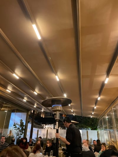This image depicts a lively evening scene within what appears to be a covered outdoor patio or veranda of a restaurant or cafe in Chaidari, Greece. The perspective is tilted, showing a section of a beige-colored ceiling or upper wall on the left, adorned with multiple horizontal light strips that cast a warm, ambient glow. These linear lights are evenly spaced, illuminating the textured surface.
In the mid-ground, a standing patio heater is prominent, emitting a visible blue flame, suggesting a cool evening temperature. A person, likely a staff member or waiter dressed in a black shirt, is seen near the heater, possibly attending to it or serving.
To the right, several patrons are seated at tables, visible through glass partitions that enclose the dining area. Their faces and figures are partially obscured, but they appear to be engaged in conversation and dining. Glasses, bottles, and other dinnerware are discernible on the tables. Beyond the glass, hints of outdoor greenery and the dimly lit exterior indicate it is nighttime. The overall atmosphere is warm and inviting, created by the artificial lighting and the gathering of people. No discernible text is visible within the frame.
