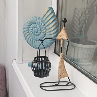 On a white windowsill, likely indoors, a light blue and gold nautilus shell sculpture stands alongside a black metal lantern and a metal sculpture of a person wrapped in brown twine. The window behind the objects reflects a potted plant. There is no indication of location within Ketoshi, Russia. No people are visible, and no actions or interactions are occurring.
