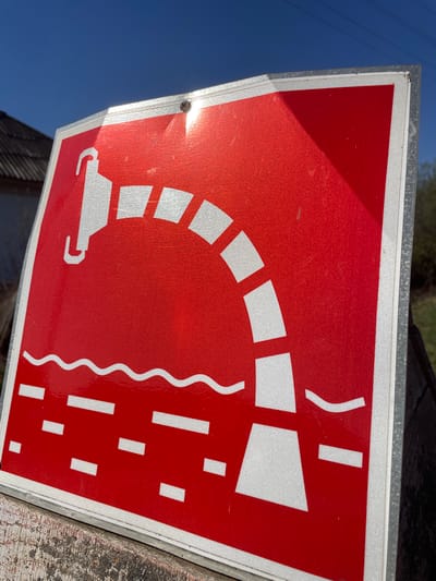 A close-up, angled shot depicts a red and white road sign. The sign, encased in a metal frame, is likely made of a reflective material given the strong sunlight reflection. The image on the sign features white symbols on a red background. The symbols depict a stylized bridge or viaduct, with a vehicle passing underneath. To the right of the bridge is a wavy vertical line, and below, a series of short, vertical lines resembling rain. The sign is mounted on a wooden surface. A portion of the clear blue sky is visible in the lower left corner of the image. The location is reported to be in Поедуги, Russia, though there are no inherent geographic markers in the image to confirm this.