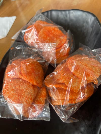 In Pulokiton, Indonesia, three clear plastic bags filled with round, orange-dusted chips are arranged on a black, textured surface, likely a bowl. The chips appear to be thin and crispy, with visible orange seasoning powder on their surfaces. The lighting suggests an indoor setting, possibly a dining area, with a wooden surface and a white napkin visible in the background. The scene is focused on the food items, with no people or discernible actions taking place.
