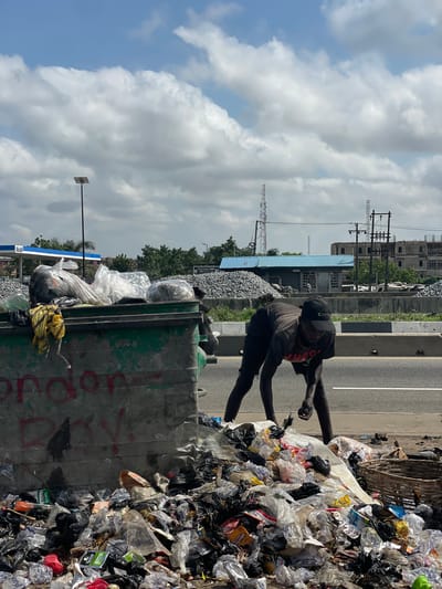 In Isunba, Nigeria, two individuals are scavenging through a large pile of trash beside a road. The trash pile contains various plastic bags and discarded items. A green dumpster is partially visible, filled with more trash. The environment includes power lines, a blue building, and a gas station in the background. The sky is cloudy.