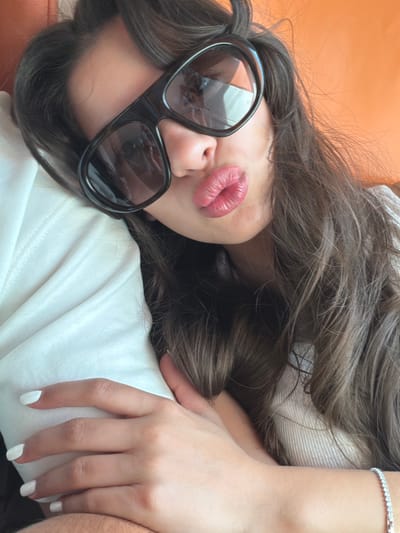 A close-up, high-angle shot captures a person, likely female, lying down in Cagnes-sur-Mer, France. Her dark, wavy hair cascades around her head, framing her face. She is wearing large, black-framed sunglasses, reflecting light and partially obscuring her eyes and upper face. Her lips are a prominent feature, colored with red or deep pink lipstick and puckered into a playful kiss pose.
She is dressed in a white garment, possibly a t-shirt or sweater, with visible fabric folds. A shimmering diamond-studded bracelet adorns her left wrist. Her left hand, with neatly painted white fingernails, rests on what appears to be another person's arm, indicated by the darker, slightly hairy skin visible at the bottom right of the frame. The background directly behind her head is a solid, warm orange color, suggesting a couch, pillow, or wall. The scene conveys a relaxed and casual moment, taken in what appears to be daytime, given the use of sunglasses. No text is visible in the image.