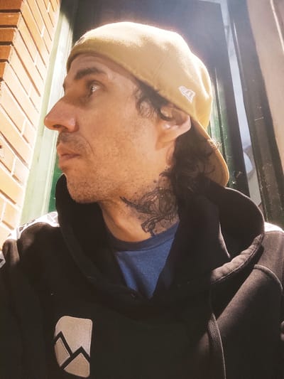 A male subject is depicted in a side profile, facing towards the left of the frame, within the city of Remedios de Escalada, Argentina. He is wearing a beige baseball cap backward, which prominently displays a white "New Era" logo on its right side. Short, dark hair is visible beneath the cap and extending onto his neck. The subject has light stubble on his face, a prominent nose, and his left ear is visible. A dark, detailed tattoo, resembling a winged creature or bird, is present on the left side of his neck. He is dressed in a dark hooded sweatshirt (hoodie) over a blue t-shirt, which is visible at the collar. The hoodie features a distinct rectangular emblem on the left chest area, characterized by a grey and beige stylized mountain or zigzag pattern. White or light-colored patterning is also visible on the shoulders or upper sleeves of the hoodie. His left eye is visible, showing a prominent highlight, and he appears to be looking upwards and to the left. To the left of the subject, a section of an orange-brown brick wall with white mortar lines is visible. To the right, behind the subject, there is a dark-framed structure, likely a window or doorway, with a dark interior or reflective surface beyond. The lighting suggests a localized light source, creating strong highlights on the subject's head and face.