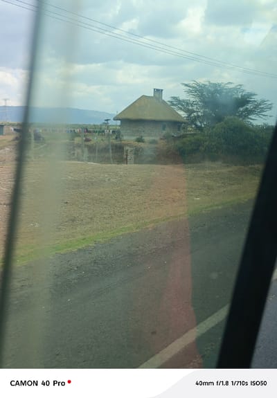 The scene depicts an outdoor rural landscape in Malewa West ward, Kenya, viewed through a reflective window. A single-story structure with stone walls, a prominent conical thatched roof, and a stone chimney is centrally positioned in the midground. Two windows are visible on its façade. To the left of this house, colorful laundry hangs on a clothesline in a dry, grassy field. This area is delineated by a rudimentary fence and two stone gateposts. Further left, a smaller building with a corrugated iron roof is partially visible. A large, spreading tree stands behind the main house, accompanied by smaller bushes. Overhead, multiple parallel utility lines traverse the sky, and a power pylon is identifiable on the far left horizon against distant hills. The foreground consists of a dark, likely gravel or asphalt road, bordered by a strip of dry and green grass. The sky is partially cloudy. No individuals are visible.