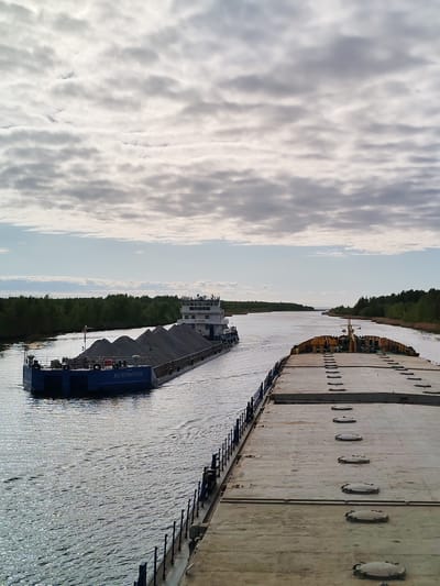 The image shows a river scene in the city of Ozernoe Ust'e, Russia. Two large barges are on the water, with the closest one taking up most of the frame. This barge has a gray, concrete-like surface with evenly spaced round hatches. A dark metal railing runs along its edge. The second barge, further away, is loaded with large mounds of gray material, possibly gravel or ore. It is being pushed by a tugboat. The river is bordered by green trees on both sides. The sky is overcast with gray and white clouds.