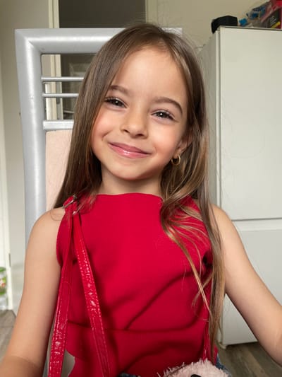 A young girl, appearing to be a child or pre-teen, is the primary subject of this close-up, brightly lit portrait. She has long, straight brown hair that cascades over her shoulders and back. She is wearing a sleeveless red top with a textured, possibly ribbed, fabric. Small, gold-colored hoop earrings are visible in her ears. A glossy red strap, potentially from a handbag or part of her outfit, crosses over her right shoulder. To the far right, a glimpse of a fluffy, light-colored object, possibly a toy or a bag detail, is visible.

The girl is smiling broadly, with a cheerful and engaging expression, looking directly at the camera. Her lips appear to have a light gloss.

The background suggests an indoor setting, likely a home in Günzburg, Germany. It features light-colored walls or furniture, possibly a cabinet or wardrobe, and a metallic, grid-like structure (which could be part of a chair, bed frame, or a vent) near the bottom left of the frame. The overall lighting suggests it is daytime, though specific time or weather cannot be determined. No visible text is present in the image.