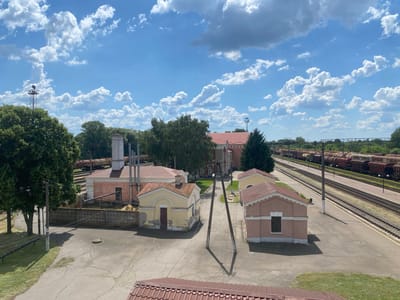 This image depicts an outdoor scene at a train station in Romodan, Ukraine, on a sunny day with a blue sky and scattered clouds. The foreground features a cracked asphalt surface with grassy patches and a red-tiled roof visible at the bottom. Several small, weathered buildings with red-tiled roofs are scattered throughout the middle ground. One yellow building has a dark door and a prominent chimney. Another pinkish-beige building has a single dark window. In the background, train tracks stretch into the distance, lined with numerous freight train cars. Trees and vegetation are visible on either side of the tracks, and a taller building with a red roof is situated further back. A few power poles with wires are also present in the scene. There are no people visible in the image.