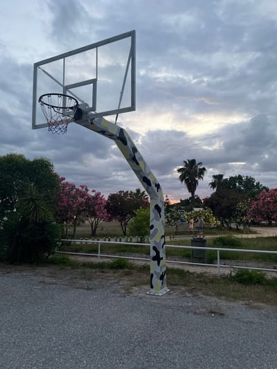 A basketball hoop with a clear backboard and a camouflage-patterned pole stands in the foreground. The hoop has a red, white, and blue net. The background features a cloudy sky, palm trees, and flowering bushes. Two people are visible in the middle ground, standing near a bench. The scene appears to be a park or recreational area in Gerakini, Greece.